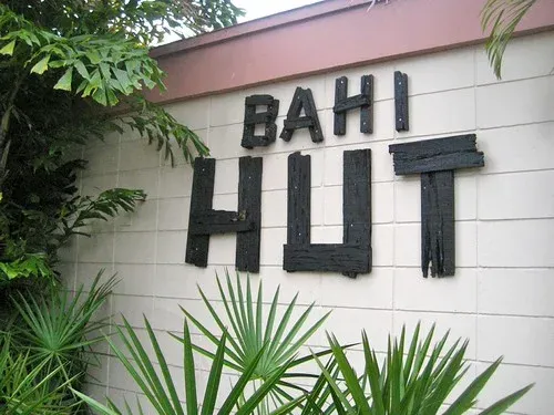 Carved wooden letters spelling BAHI HUT on a white block wall, framed by palm fronds